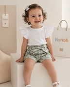 Conjunto Pérolla – Menina sentada sorrindo com conjunto branco e shorts floral